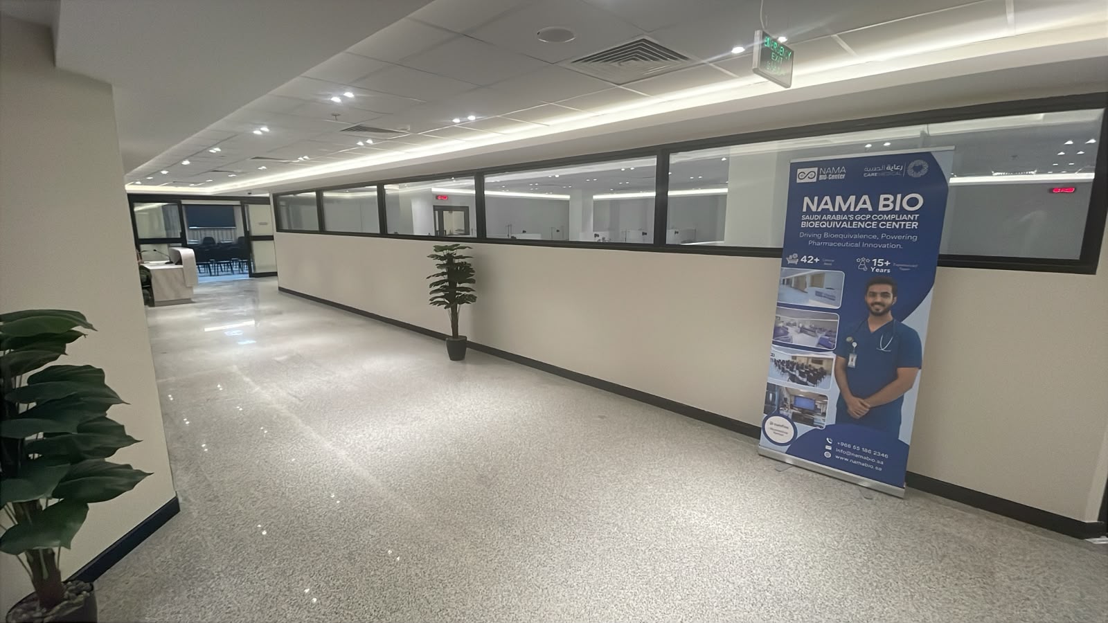 A wide, bright corridor at NAMA Bio-Center running alongside the clinical ward, separated by a long band of internal observation windows, with a NAMA BIO roll-up banner at the far end.