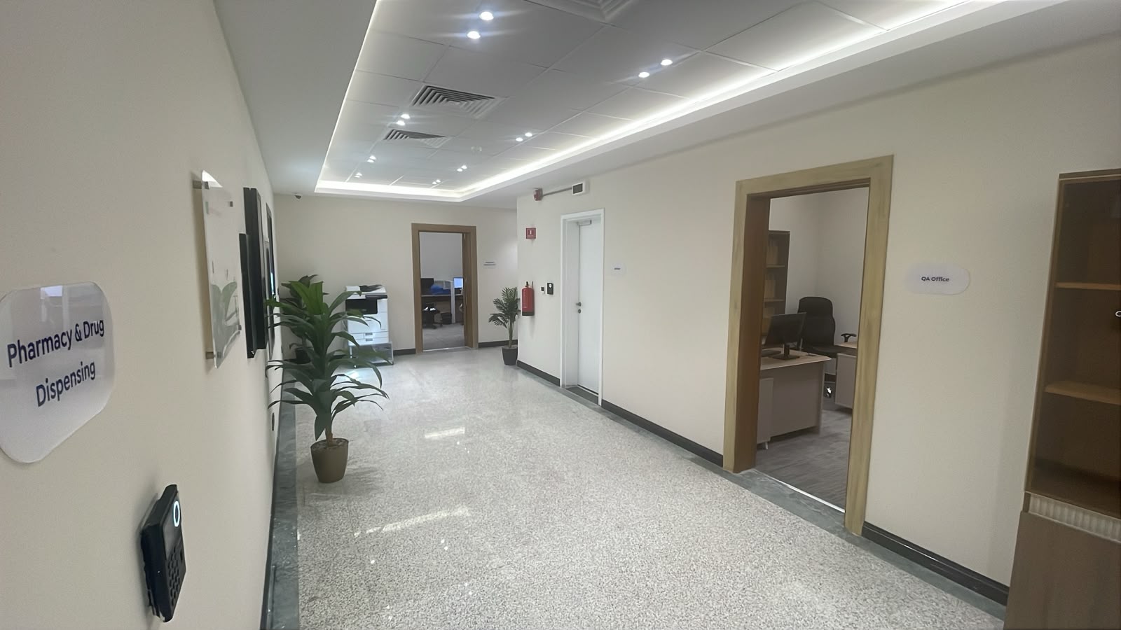 A bright corridor at NAMA Bio-Center with door signage reading Pharmacy & Drug Dispensing and QA Office, with planting and a printing station.