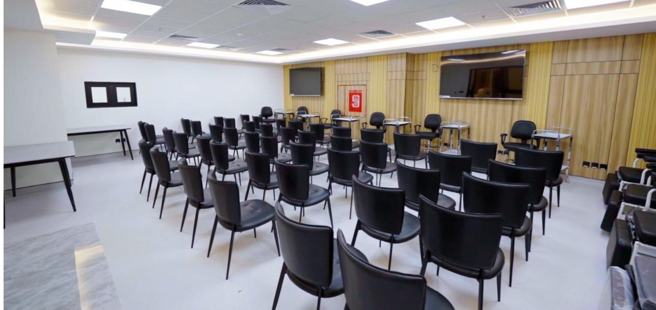 The NAMA Bio-Center training room, arranged auditorium-style with rows of chairs facing two wall-mounted screens on a wood-panelled wall.